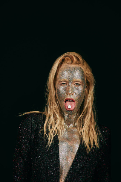 προκλητική νεαρή ξανθιά γυναίκα με glitter μακιγιάζ δείχνει γλώσσα με χάπι σε μαύρο φόντο - Φωτογραφία, εικόνα