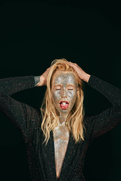 νεαρή ξανθιά γυναίκα με glitter μακιγιάζ και κλειστά μάτια δείχνει γλώσσα με χάπι σε μαύρο φόντο - Φωτογραφία, εικόνα