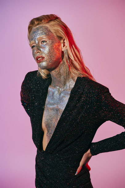 mulher loira impressionante com maquiagem brilho e vestido brilhante posando com as mãos sobre os quadris em pano de fundo rosa - Foto, Imagem