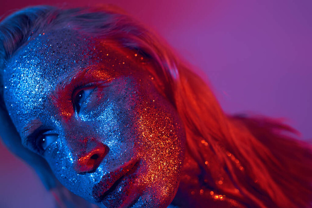 close-up de mulher jovem e loira com brilho cintilante no rosto posando no fundo de néon vibrante - Foto, Imagem