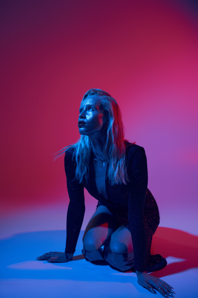 blonde and stylish  woman covered in glitter posing in shiny outfit under colored lights in studio - Photo, Image