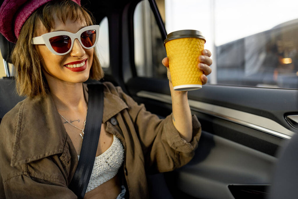 Junge, stylische Frau mit Kaffeetasse auf dem Rücksitz eines modernen Taxis während der Fahrt in der Stadt. Kaffeepause für unterwegs - Foto, Bild
