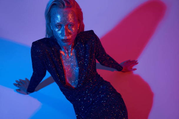 alluring sparkling model covered in glitter posing in shiny outfit under colored lights in studio - Photo, Image