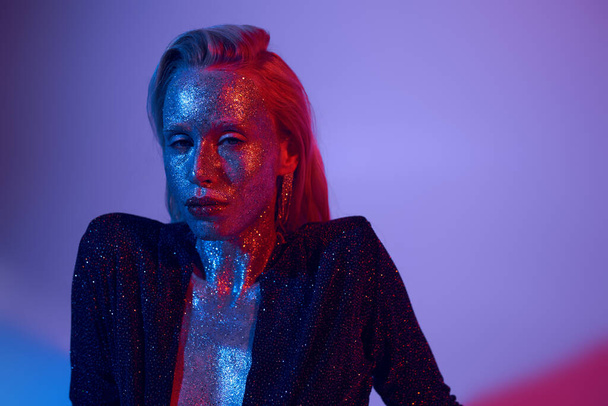 portrait of sparkling woman covered in glitter posing in shiny outfit under colored lights in studio - Photo, Image