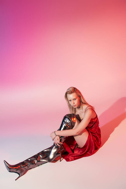 beautiful stylish woman with long blonde hair in red dress sitting on floor on pink background - Photo, Image