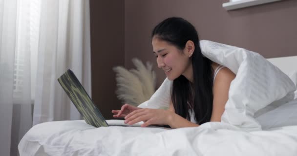 Una giovane donna di bellezza asiatica si trova sul letto coperto da una trapunta e naviga siti web su laptop, negozi online, chat con gli amici. - Filmati, video