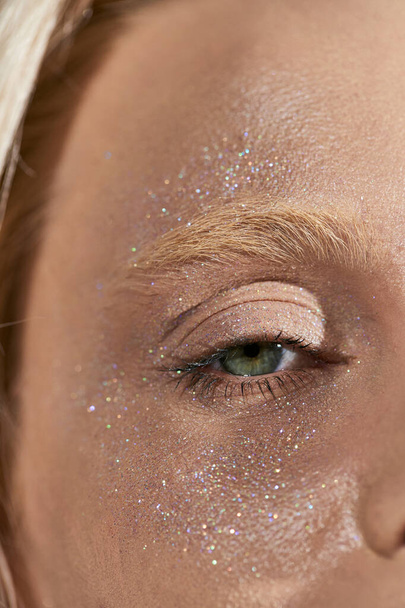 κοντινό πλάνο της νεαρής γυναίκας με πράσινο μάτι και λαμπερό glitter στο πρόσωπό της κοιτάζοντας κάμερα, highlighter - Φωτογραφία, εικόνα