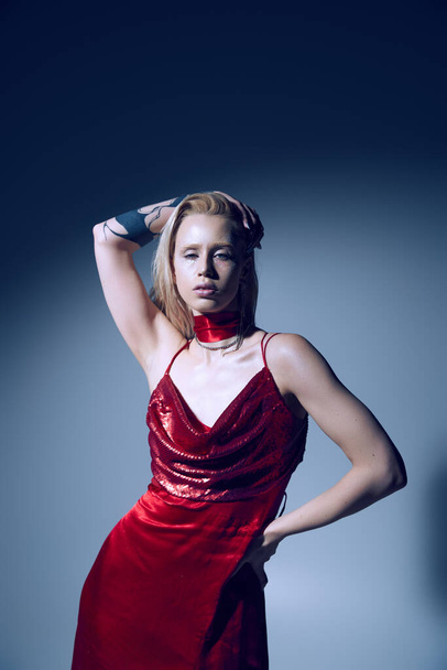 mulher sexy elegante com tatuagem e iluminador em vestido vermelho com um braço levantado e outro na anca - Foto, Imagem
