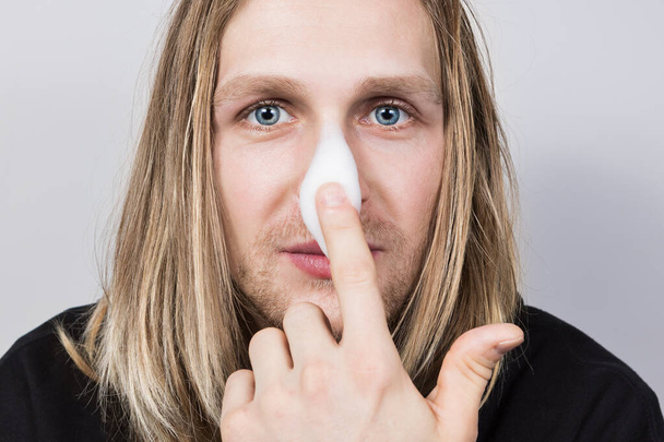 A real young handsome long haired blond with blue eyes and applying cleansing foam on his nose with the finger of one hand.Cosmetology for men,professional and home skin and hair care,closeup - Photo, Image