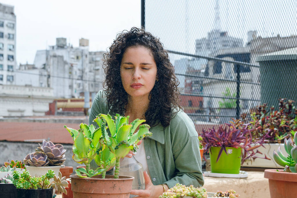 junge lateinische Frau in Lockenwicklern, bei der Arbeit stehend, Pflanzen auf der Terrasse wartend, Wassersprüher benutzend, um sie zu hydratisieren, Heimarbeitskonzept, Kopierraum. - Foto, Bild