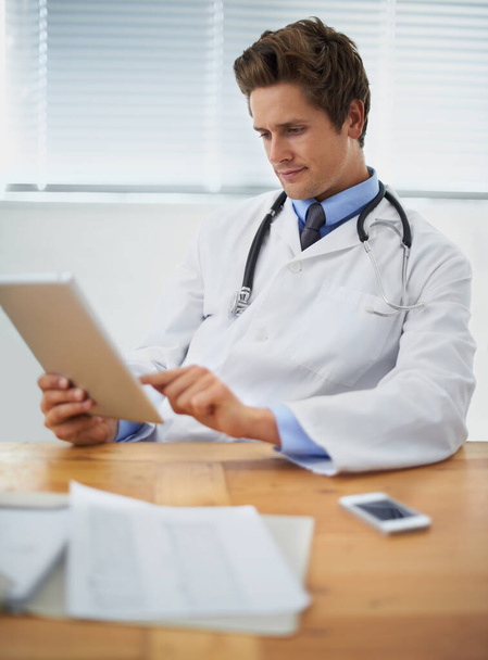 Tablet, research and doctor reading in his office for diagnosis or treatment at medical hospital. Internet, digital technology and professional young male healthcare worker sitting by desk in clinic - Photo, Image