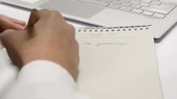 Close-up van een zakenman, zijn gezicht meestal verborgen, schrijven op een notitieblok voor het typen op een laptop computer in een helder modern stadskantoor. - Video