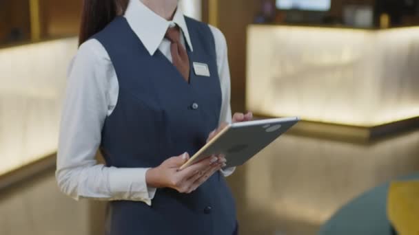 Tilt up πλάνο της γυναίκας διευθυντής του ξενοδοχείου με επίσημη στολή στέκεται στην αίθουσα εισόδου και χρησιμοποιώντας ψηφιακή ταμπλέτα - Πλάνα, βίντεο