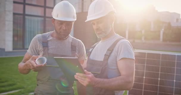 Two multiethnic engineers working at solar panel station. African American worker holding drawing. Caucasian technician man holding laptop. Two multicultural contractors installing solar panels. - Footage, Video
