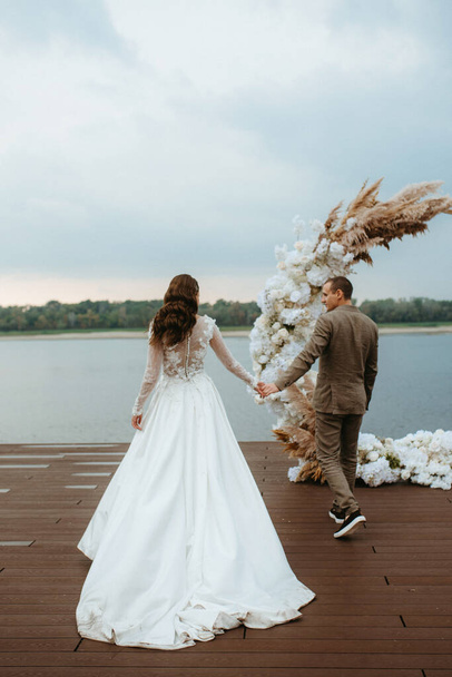 der erste Hochzeitstanz des Brautpaares auf der Seebrücke am Fluss, vor dem Hintergrund des Hochzeitsbogens - Foto, Bild
