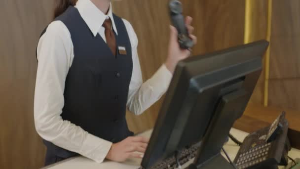 Νεαρή γυναίκα διευθυντής ρεσεψιόν σε formalwear σηκώνοντας το τηλέφωνο, μιλώντας με τον πελάτη και χρησιμοποιώντας υπολογιστή κατά τη διάρκεια της εργάσιμης ημέρας - Πλάνα, βίντεο