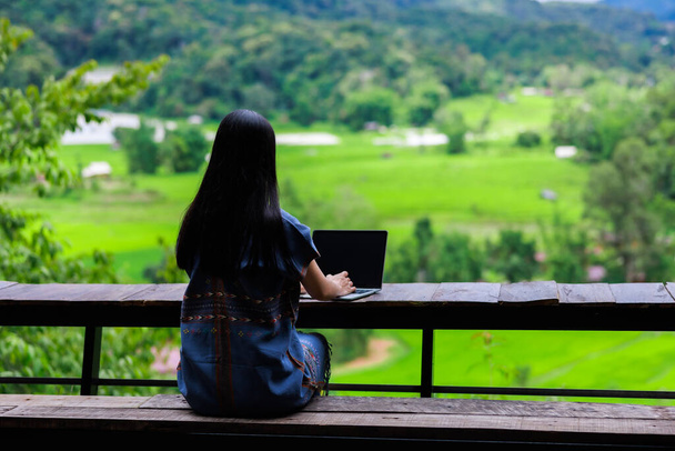 молодая женщина работает за ноутбуком, сидя на деревянном балконе с прекрасным видом на природу,  - Фото, изображение