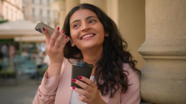 Joven sonriente feliz mujer de negocios india árabe mujer étnica chica mujer trabajadora de negocios escuchando mensaje de voz teléfono inteligente beber taza de café recibir audio utilizando los medios sociales de telefonía celular fuera de la ciudad - Imágenes, Vídeo