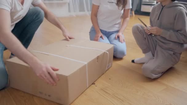 Мальчик и его родители, сидя на полу, с нетерпением распаковывают большую картонную коробку из посылки с товарами, которые были доставлены в их дом. Покупки онлайн с заказами на доставку. - Кадры, видео