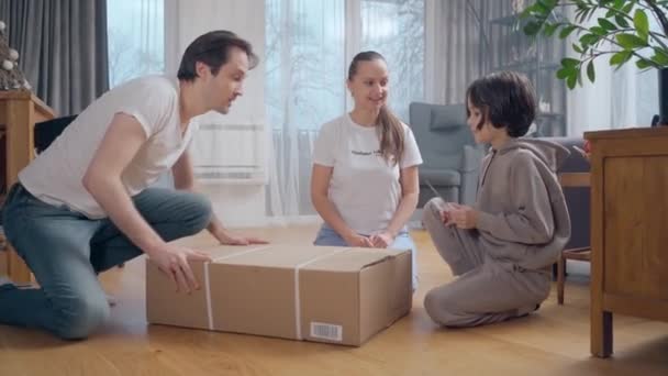 De jongen en zijn ouders, die thuis op de grond zaten, pakten ongeduldig een grote kartonnen doos uit met een pakje goederen dat bij hen thuis werd afgeleverd. Familie winkelen online met levering aan huis. - Video