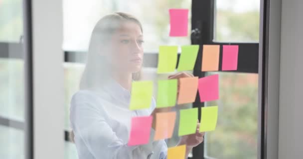 Geconcentreerde vrouw organiseert gedachten door kleurige stickers op glasplaat te plakken. Zakenvrouw gebruikt stickers om belangrijke punten te markeren tijdens vergadering - Video