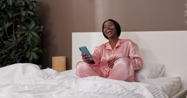 Nuori nainen istuu sängyllä katsomassa hassuja videoita puhelimestaan. Käsite käyttää älypuhelinta heräämisen jälkeen. - Materiaali, video