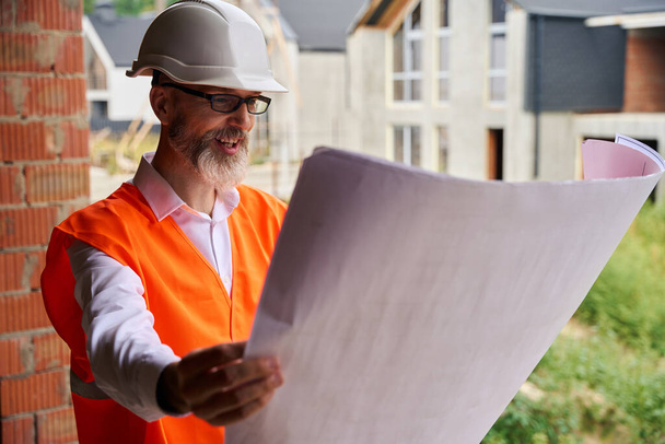 Waist-up portrait of contented foreman looking at blueprints in his hands while standing among half-built houses - 写真・画像
