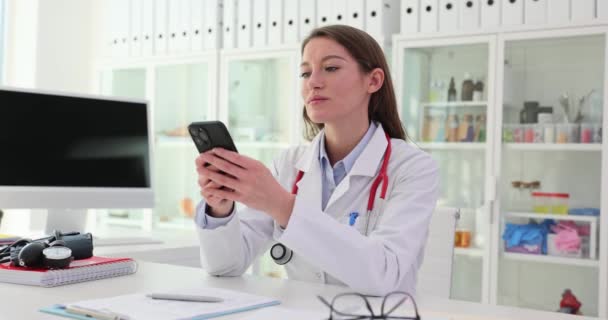Woman physician sits at desk and types message on smartphone. Family doctor drafts notification on modern device to inform patient about appointment - Footage, Video