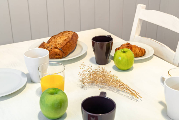 Свежий вкусный завтрак подается на белом столе с круассанами, яблоками и апельсиновым соком - Фото, изображение