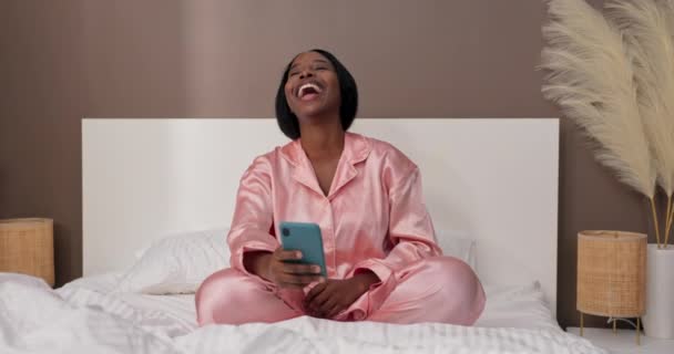 Uma jovem se senta na cama assistindo vídeos engraçados no telefone. O conceito de usar um smartphone depois de acordar. - Filmagem, Vídeo