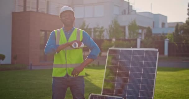 Férfi vállalkozó tükröződő mellényben és mosollyal. Afrikai amerikai mérnökök rajzzal a kezükben pózolnak. Vállalkozó keménykalapban, napelem mellett áll. A napfény villamos energiává alakítása. - Felvétel, videó