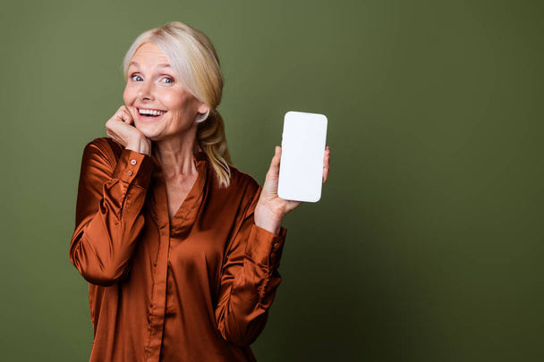 Portrait of impressed person with blond hair wear blouse presenting online store on smartphone display isolated on khaki color background. - Foto, Imagen