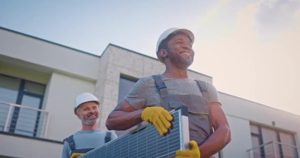 Dos sonrientes ingenieros multiétnicos instalando una fuente de energía alternativa. Contratistas masculinos hablando y llevando panel solar. Dos hombres multiculturales que trabajan en la empresa de energía verde. - Metraje, vídeo