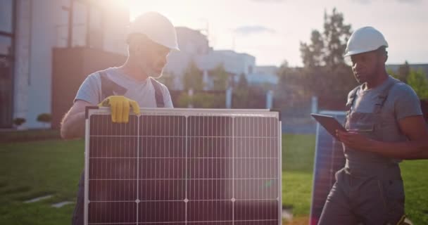 Два мультикультурных человека работают на предприятии возобновляемых источников энергии. Многонациональные подрядчики стоят на газоне. Инженеры-технологи устанавливают солнечные панели. Афроамериканец держит планшет. - Кадры, видео