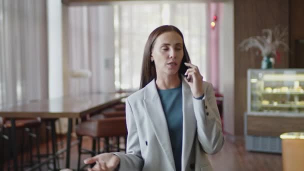 Medium shot van jonge zakenvrouw in formele pak lopen door hotel restaurant en het hebben van gesprek op mobiele telefoon - Video