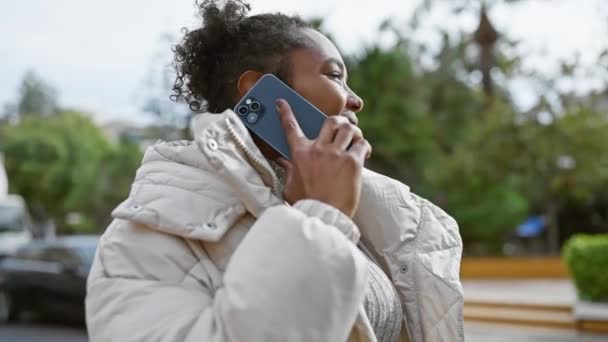 Egy göndör hajú, mosolygós fiatal nő beszél okostelefonon, miközben egy városi utcán áll, megtestesítve a modern városi életet.. - Felvétel, videó