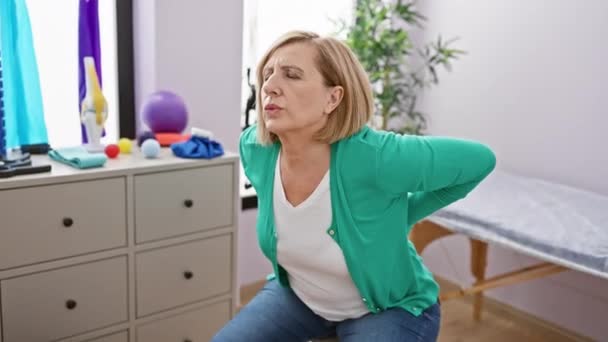 Mulher branca madura com dor em uma clínica de fisioterapia dentro de casa, sentada e tocando suas costas. - Filmagem, Vídeo