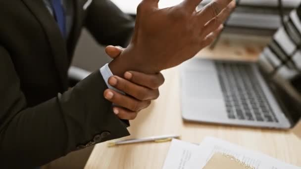 Ein professioneller schwarzer Mann untersucht seine Schmerzen am Handgelenk in einem Büro mit Laptop und Dokumenten auf dem Schreibtisch. - Filmmaterial, Video