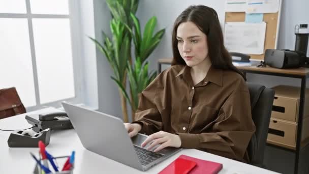 Una joven sorprendida trabajando en su portátil en una moderna sala de oficina, reflejando un escenario profesional en interiores. - Metraje, vídeo