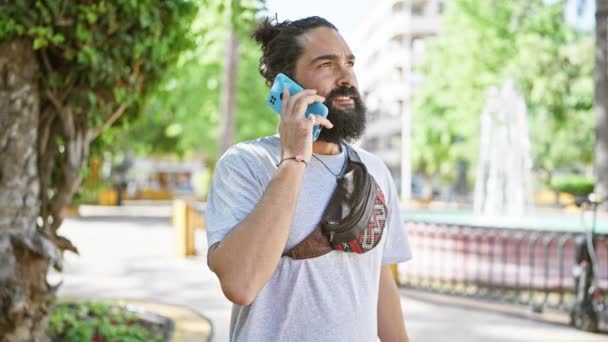 Uśmiechnięty brodaty mężczyzna trzymający smartfona mówi i pisze podczas spaceru po słonecznej ulicy miasta. - Materiał filmowy, wideo