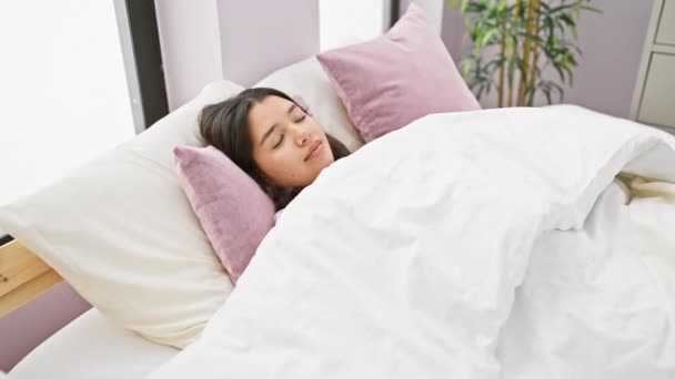 Eine junge hispanische Frau, die sich mit einem Lächeln im Bett ausbreitet und eine gemütliche Morgenroutine in einem gut beleuchteten Schlafzimmer darstellt. - Filmmaterial, Video