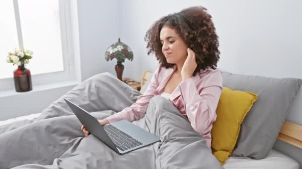 Eine junge Frau mit lockigem Haar benutzt einen Laptop, während sie es sich in einem gemütlichen Schlafzimmerinnenraum gemütlich macht und einen lässigen und komfortablen Lebensstil ausstrahlt. - Filmmaterial, Video