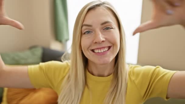 Fröhliche junge blonde Frau zu Hause, die ein T-Shirt trägt, mit ihren Fingern eine Herzform schafft, Augen voller Liebe und Glück lächelt und in einer romantischen Geste warme Gefühle ausdrückt. - Filmmaterial, Video