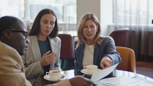 Two Caucasian female colleagues having coffee and discussing documents with African American business partner during meeting in hotel restaurant - Footage, Video