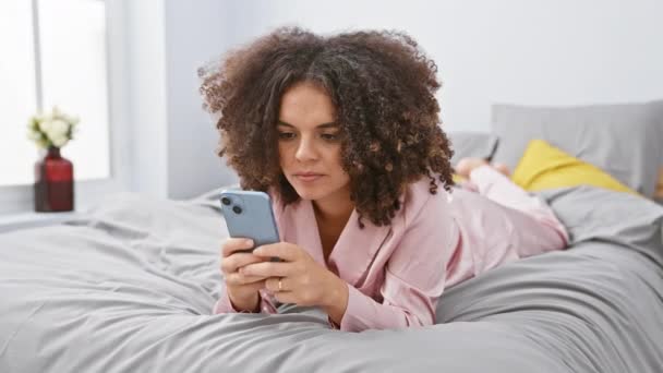 Cheerful hispanic woman with curly hair lies in bed, giving thumbs-up sign of approval while texting on smartphone - Footage, Video