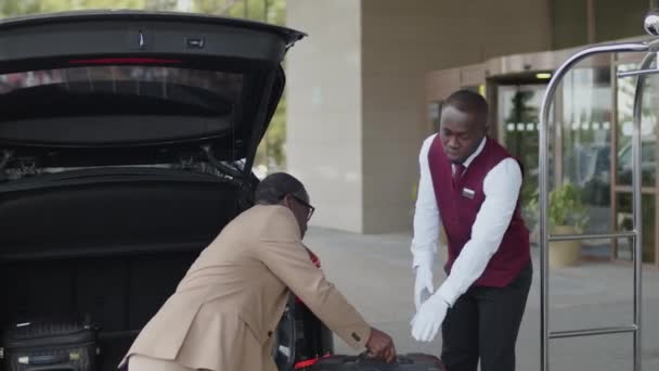 Jovem paquete afro-americano de uniforme ajudando o hóspede a descarregar malas do porta-malas do carro e colocá-las no carrinho de bagagem em frente à entrada do hotel - Filmagem, Vídeo