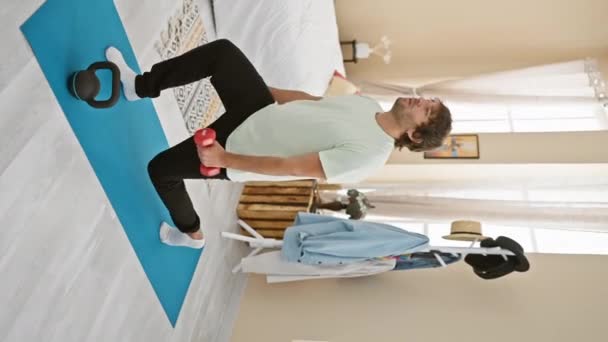 Een bebaarde jongeman oefent met halters op een yogamat in een nette slaapkameromgeving. - Video