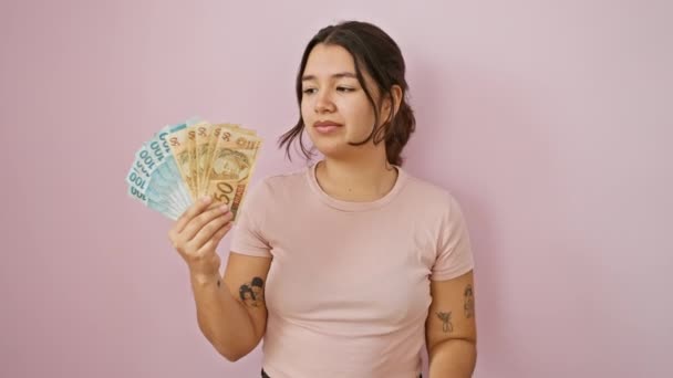 Захоплена молода іспаномовна жінка, вказуючи пальцем на бразильські справжні банкноти з допитливим виглядом, випромінює впевненість над рожевим ізольованим фоном - Кадри, відео