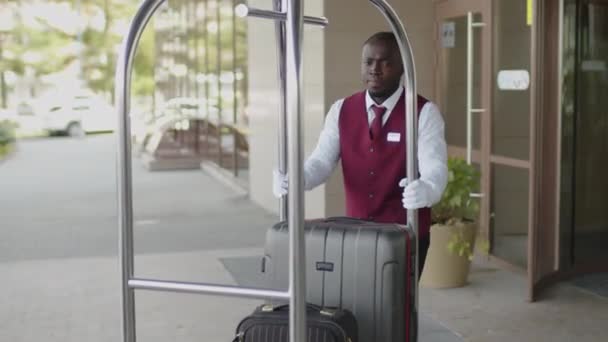 Young African American bellboy in uniform carrying suitcases on luggage cart outside of hotel - Footage, Video