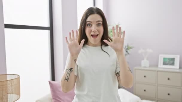 Wesoła młoda kobieta w piżamie zabawnie pokazuje znak "10" palcami siedząc na łóżku, promieniując pozytywnością w przytulnej sypialni - Materiał filmowy, wideo